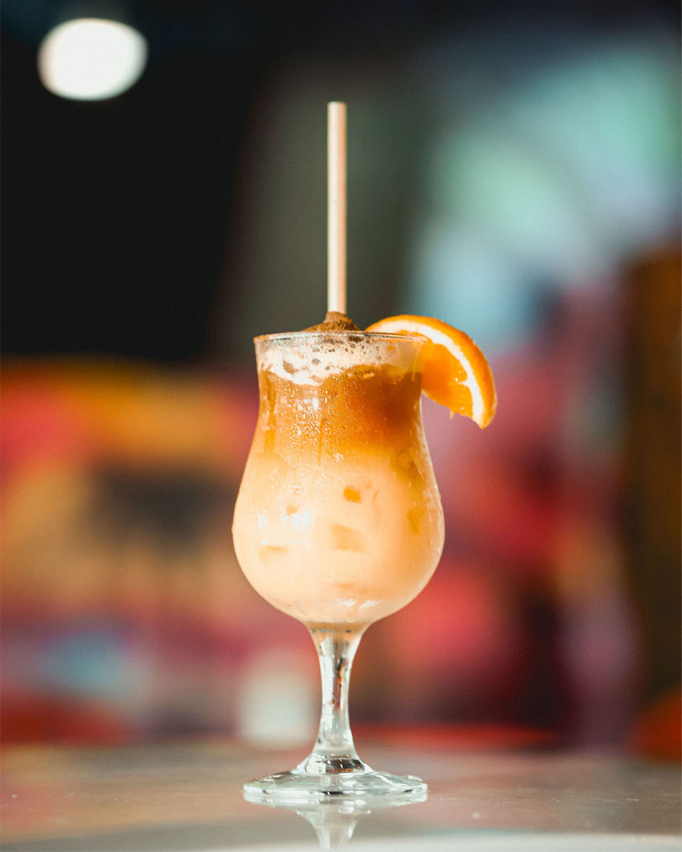 Shot of a single cocktail with a straw and orange slice on the rim, placed on top of a table.