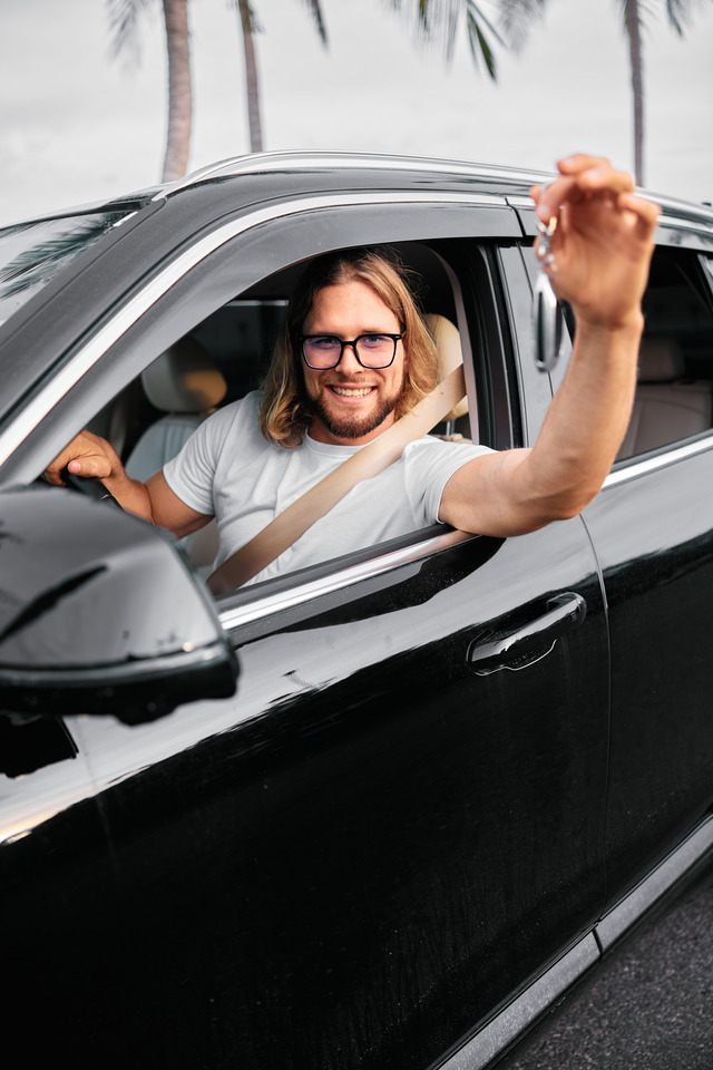 Man smiling while driving a black car, holding the keys outside the window.